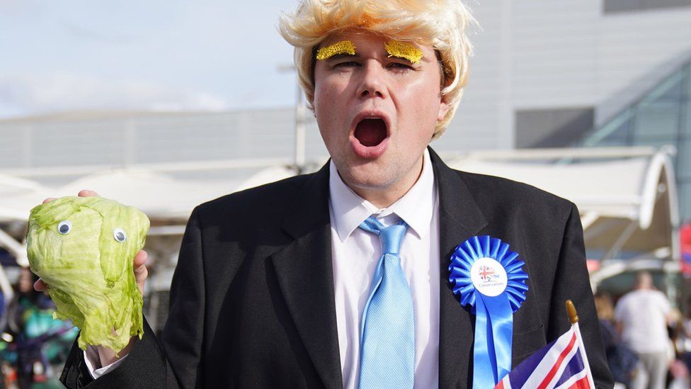 A person wearing a Boris Johnson costume with stick-on eyebrows holds a lettuce with googly eyes