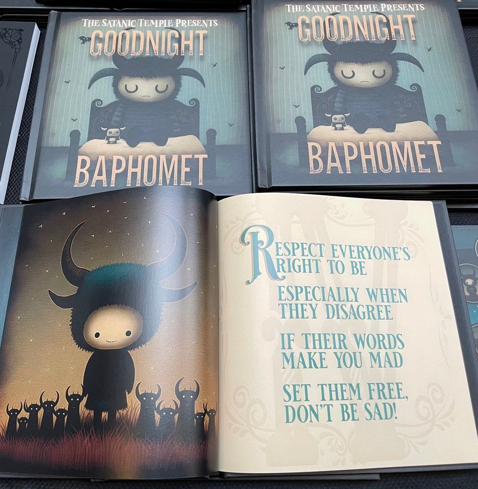 The front cover of the children's book 'Goodnight Baphomet' from The Satanic Temple shows a sleepy child baphomet - a little goat figure with horns, closing its eyes on a bed, a small toy nearby. An inside page has the rhyme: "Respect everyone's right to be, especially when they disagree. If their words make you mad, set them free - don't be sad!"