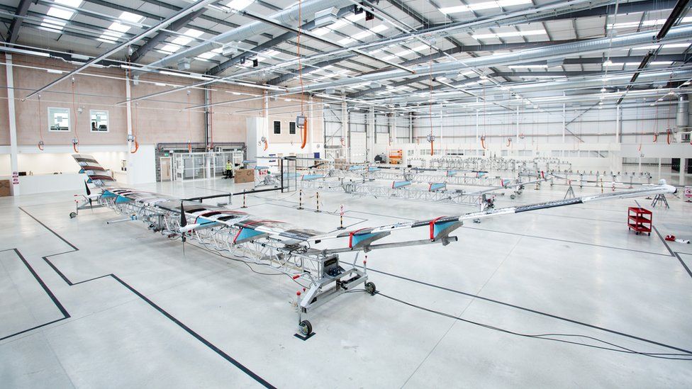 A manufacturing floor with a long, transparent glider-like plane visible in the foreground