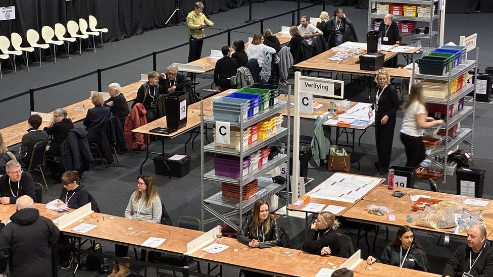 People counting votes at long wooden tables in a conference centre