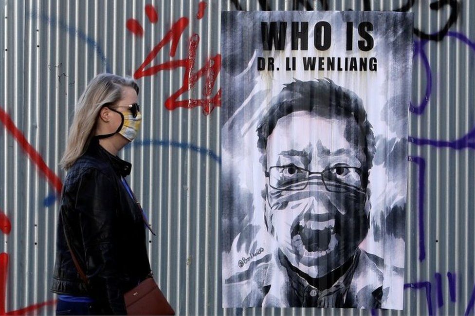Женщина в маске проходит мимо плаката с изображением покойного Ли Вэньляна в Праге