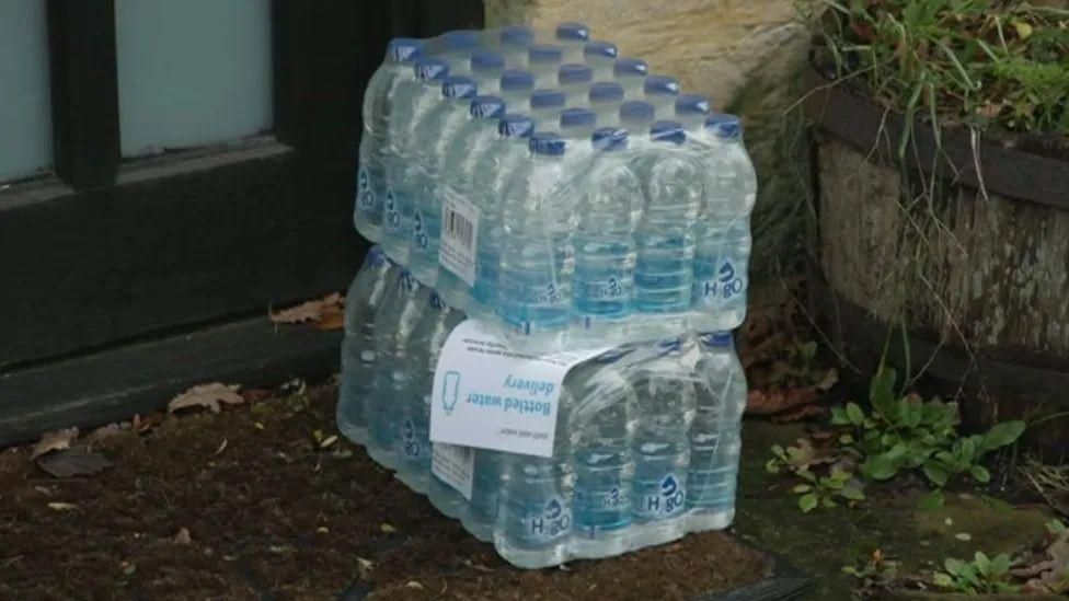 Two crates of bottled water outside a brown door. Each crate contains 24 bottles. There is a white leaflet in between the crates, however the writing is unclear