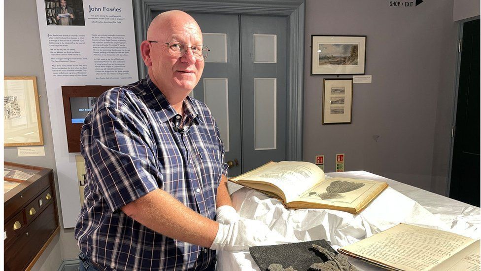 Dr Paul Davis, a middle-aged man wearing glasses and white gloves, stands next to the Mary Anning book which is on a table