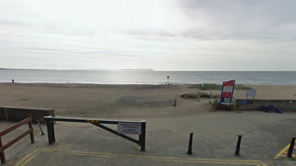 Gate and bollards at entrance to sandy beach with beach warning sign by low wall