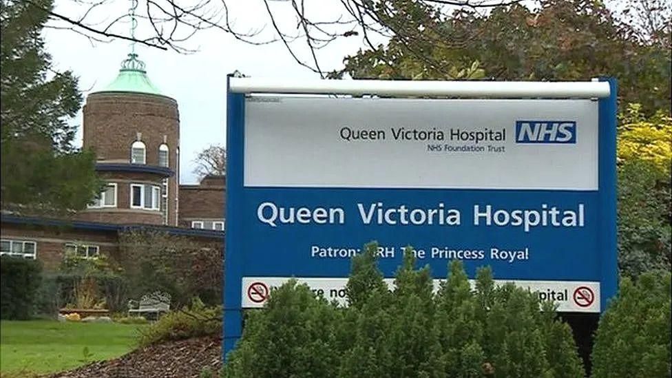 A blue and white sign reading The Queen Victoria Hospital NHS Foundation Trust. It is in front of a brown building surrounded by grass and trees