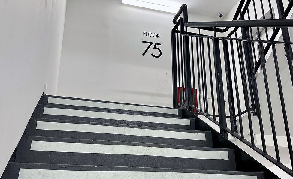 A view of the staircase at the Landmark Pinnacle building with a sign on the wall at the top of the stairs saying 'floor 75', the stairs are black and white with metallic bannisters and the walls are white.