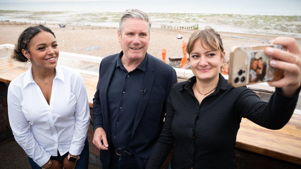 Sir Keir Starmer, the leader of the Labour Party, takes a selfie on the Worthing sea front with two young women. Sir Keir, a white man in his 60s with greying hair, wears a dark blue shirt and suit jacket. He stands between two young women, one of whom is holding her iPhone aloft to pose for the selfie she is taking. The woman on Sir Keir's right smiles at the phone and wears a white shirt and has her dark curly hair tied back. On Sir Keir's right, the young woman wears a black short and has her blonde hair tied back. Behind them is the sandy beach and sea which looks green on an overcast day.