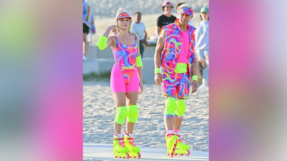 Margot Robbie as Barbie and Ryan Gosling as Ken while they were filming scenes for the Barbie move in June 2022. Both are standing on a sandy beach and both are wearing fluorescent yellow and pink outfits that are extremely bright as well as similarly coloured visors on their heads. Both also have bright yellow elbow and knee pads on and are wearing bright yellow and pink rollerblades.