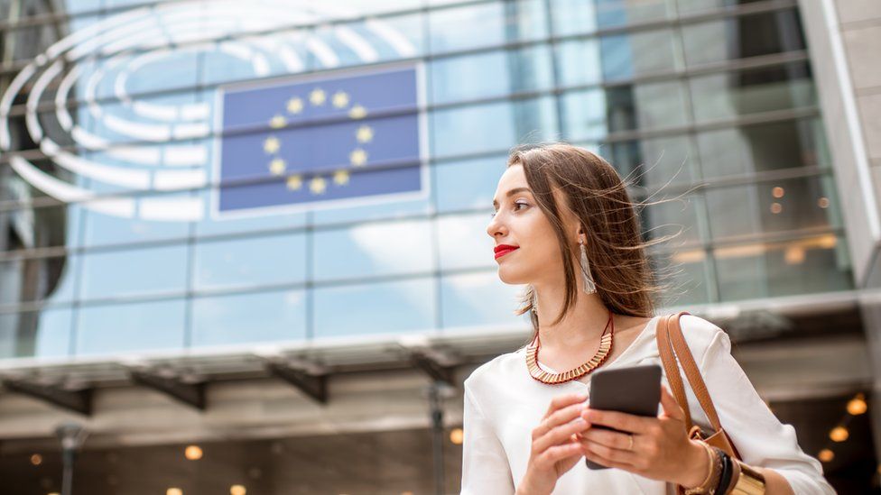 A stock image of a woman holding a phone in front of the EU parliament building