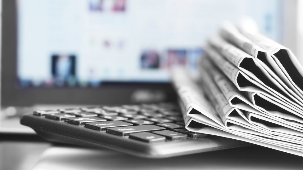 A stack of folded newspapers sits on a keyboard in front of a monitor showing a blurred news website