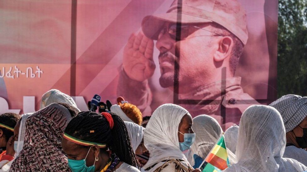 A poster with Prime Minister Abiy Ahmed wearing army uniform. There are people walking past it, one with an Ethiopian flag.