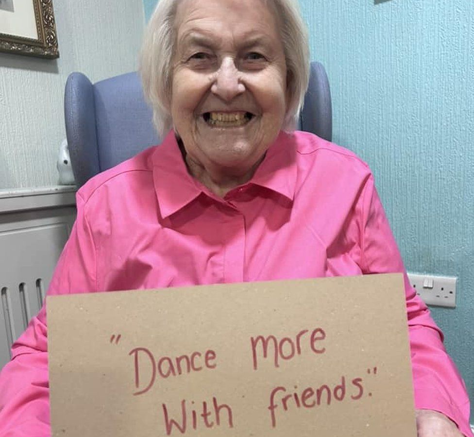 A care home resident holds a sign reading 'Dance more with friends'