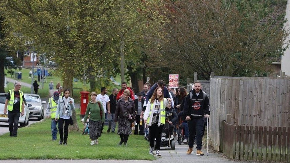 People of all ages march through a housing estate. One person is using a wheelchair, another has a stick.