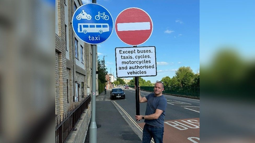 A man holds a home made "no entry" sign next to a blue street sign