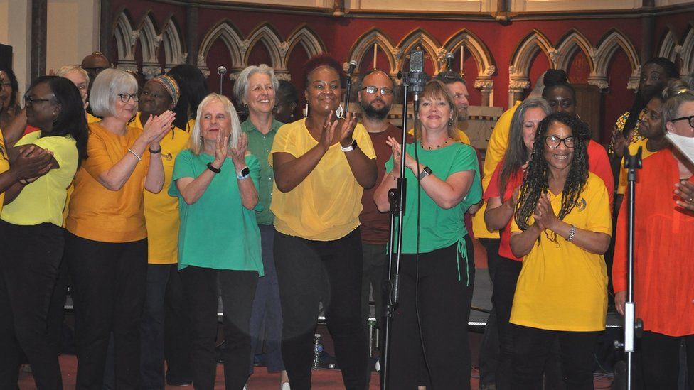 Choir members in colourful tops applaud on a stage