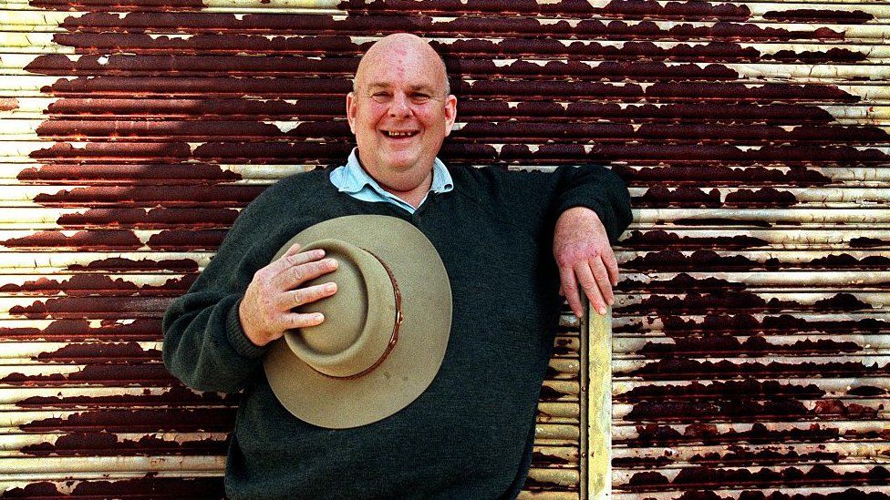 Les Murray stands holding an Akubra hat against a rusted wall