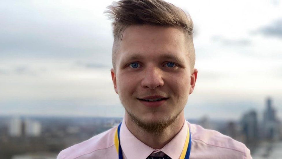 Taylor is a white man in his 20s with fair hair cut short on the sides of his head and longer on top brushed to the right. He has blue eyes and blonde stubble and wears a pale pink shirt and burgundy tie with a yellow and blue striped lanyard around his neck. He is photographed outside on an overcast day facing the camera and is smiling.