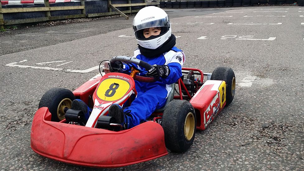 Chloe as a child, sitting in a red kart which has the race number "8" on the front in black on a yellow background. Chloe is wearing a blue race suit with white shoulders, and a white helmet, with her hands on the steering wheel. The background is the race tarmac.