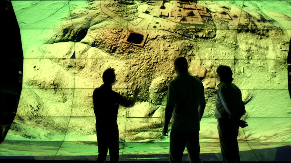 Three surveyors looks at a large digital screen showing a Lidar image of a Mayan city.