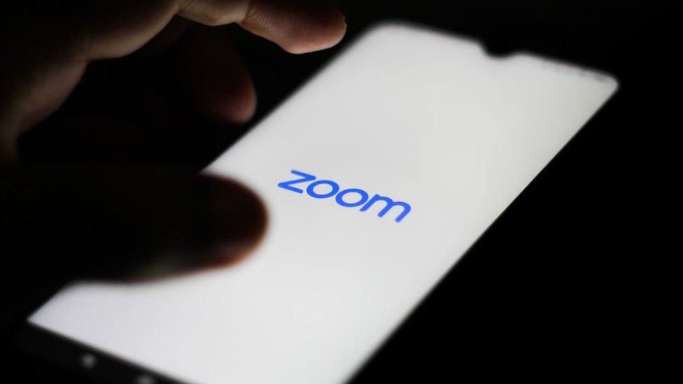 The Zoom logo is shown on a smartphone in close-up with a hand hovering nearby
