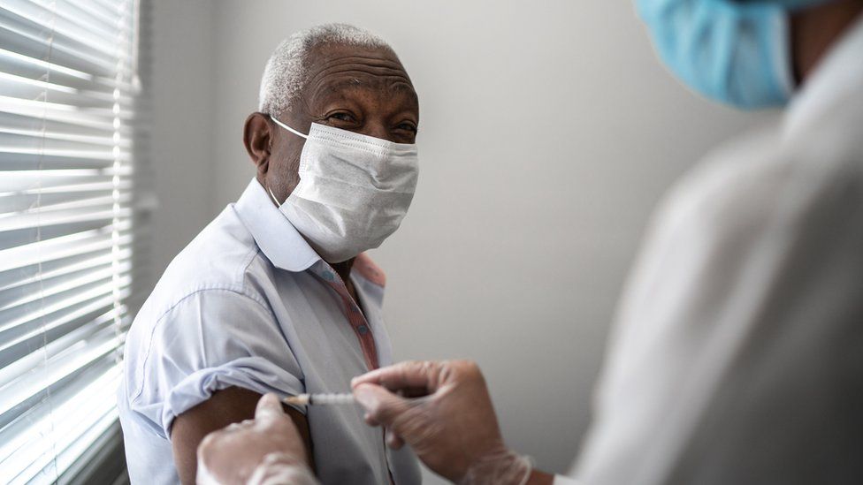Elderly man receiving vaccine on the arm, whilst wearing a face mask.