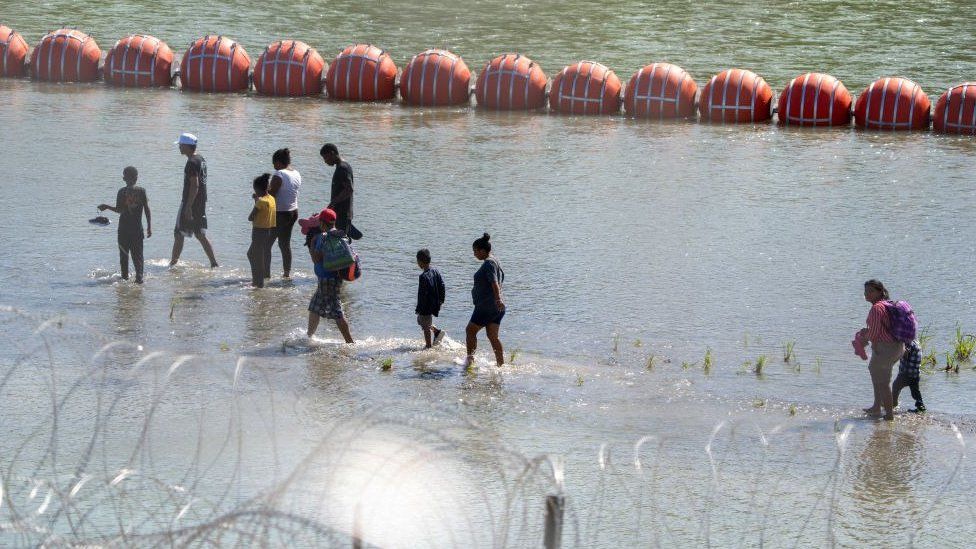 Мигранты проходят мимо буев, установленных на воде вдоль границы Рио-Гранде с Мексикой в ​​Игл-Пасс, штат Техас, 15 июля 2023 года, чтобы предотвратить въезд нелегальных иммигрантов в США.