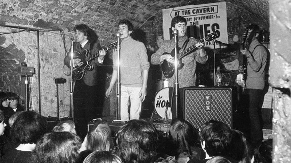 Black and white image of the band The Hollies playing on stage at The Cavern Club in front of a crowd of young people