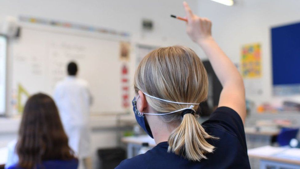 A pupil wearing a mask raises her hand in a classroom