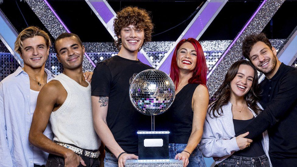 Nikita Kuzmin, Layton Williams, Bobby Brazier, Dianne Buswell, Ellie Leach & Vito Coppola. The 2023 Strictly Come Dancing finalists stand in a line with the glitterball trophy in the middle. Nikita has floppy blonde hair and wears an unbuttoned blue shirt over a white vest. He rests his hand on Layton's left shoulder. Layton, who has short dark hair, wears a white sweater vest and has his right hand in his pocket. Bobby, stands about a head taller than the rest of his cast members and has curly brown hair. He smiles at the camera, wearing a black T-shirt exposing a tattoo on his right upper arm. His partner Dianne stands next to him, she has long bright red hair and wears red lipstick and a black vest. Next in the line is Ellie, who is the shortest of the finalists, she has long brown hair and wears an unbuttoned pale shirt over a black vest. Her partner Vito is hugging her and resting his cheek on the top of her head. Vito wears a long sleeved navy blue shirt and has short brown hair.