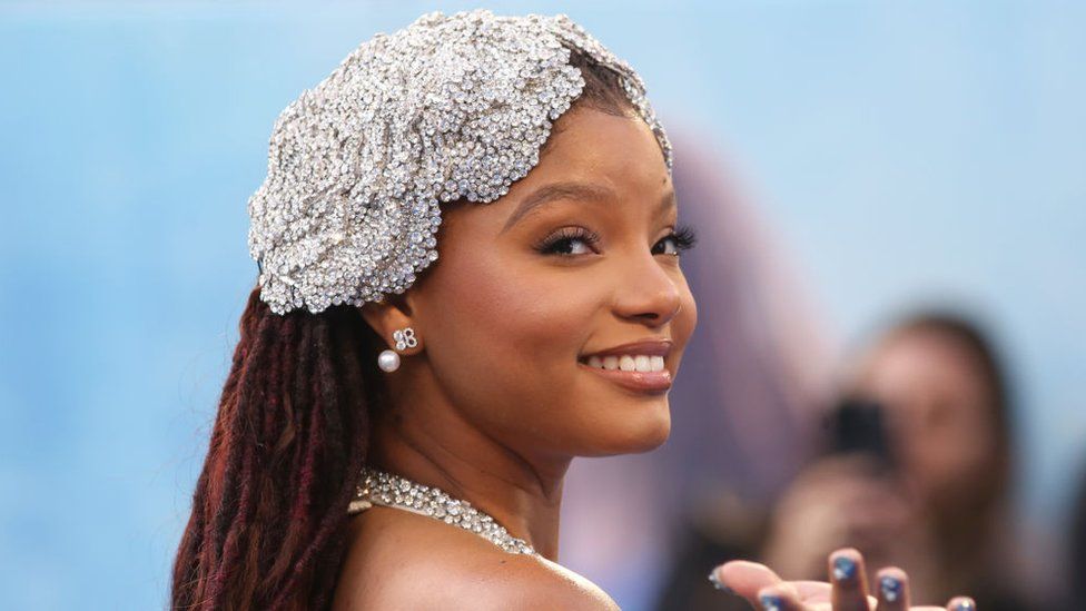 Halle Bailey on the red carpet - she's looking back over her right shoulder with a beaming smile. She's wearing a jewelled headpiece that covers the top of her head to the middle of her ear. She wears a sparkly earring with a pearl dangling from it.