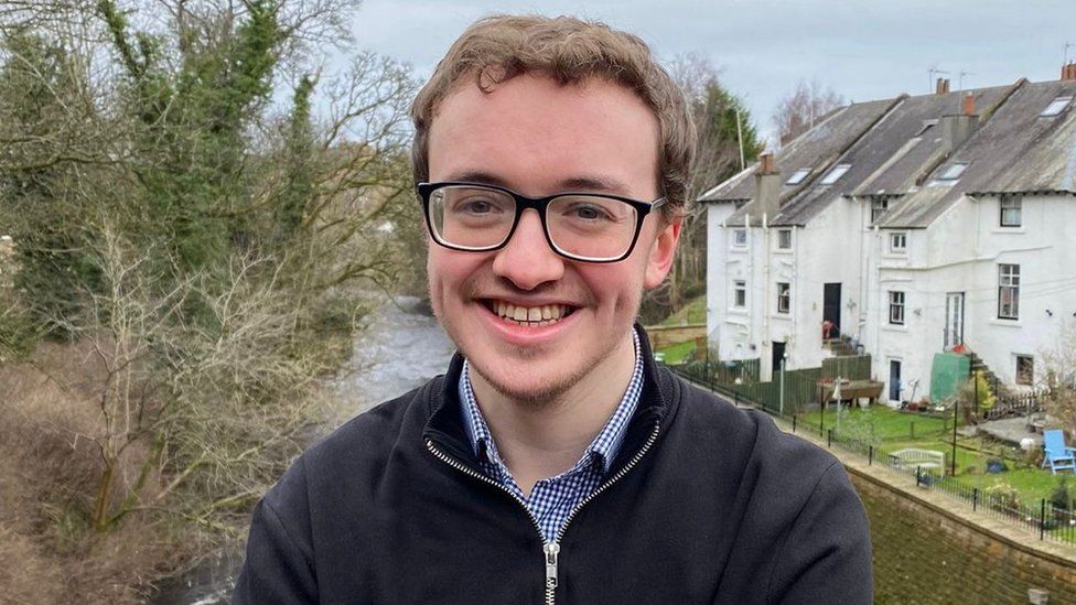 Euan Blockley smiling for the camera with a river and houses behind him. He is wearing a black fleece and has glasses