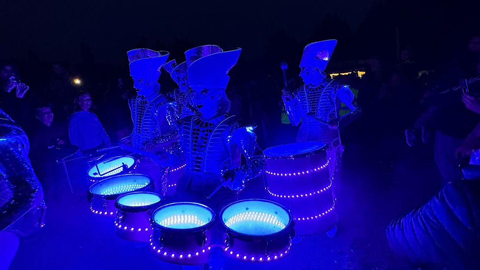 Four drummers wearing extravagant costumes carry their instruments and entertain crowds while covered in blue LEDs