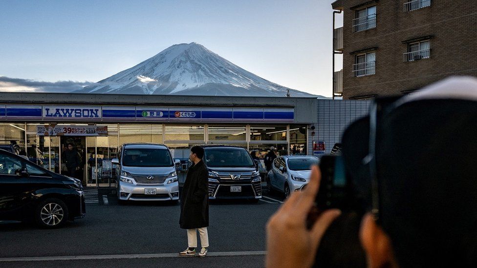 An image showing a photo being taken of a person standing in front of a Lawson shop, with Mount Fuji in the background