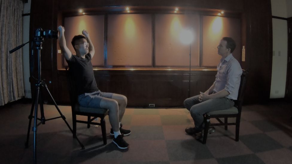 Mr Cheng, sitting down during a BBC interview, raises his arms above his head to demonstrate the position he was forced into