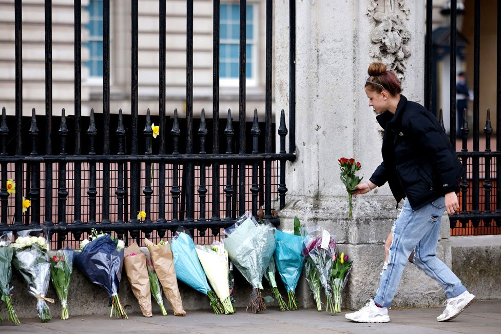 A woman adds a bunch of flowers to a line of floral tributes against the railings at the front of Buckingham Palace