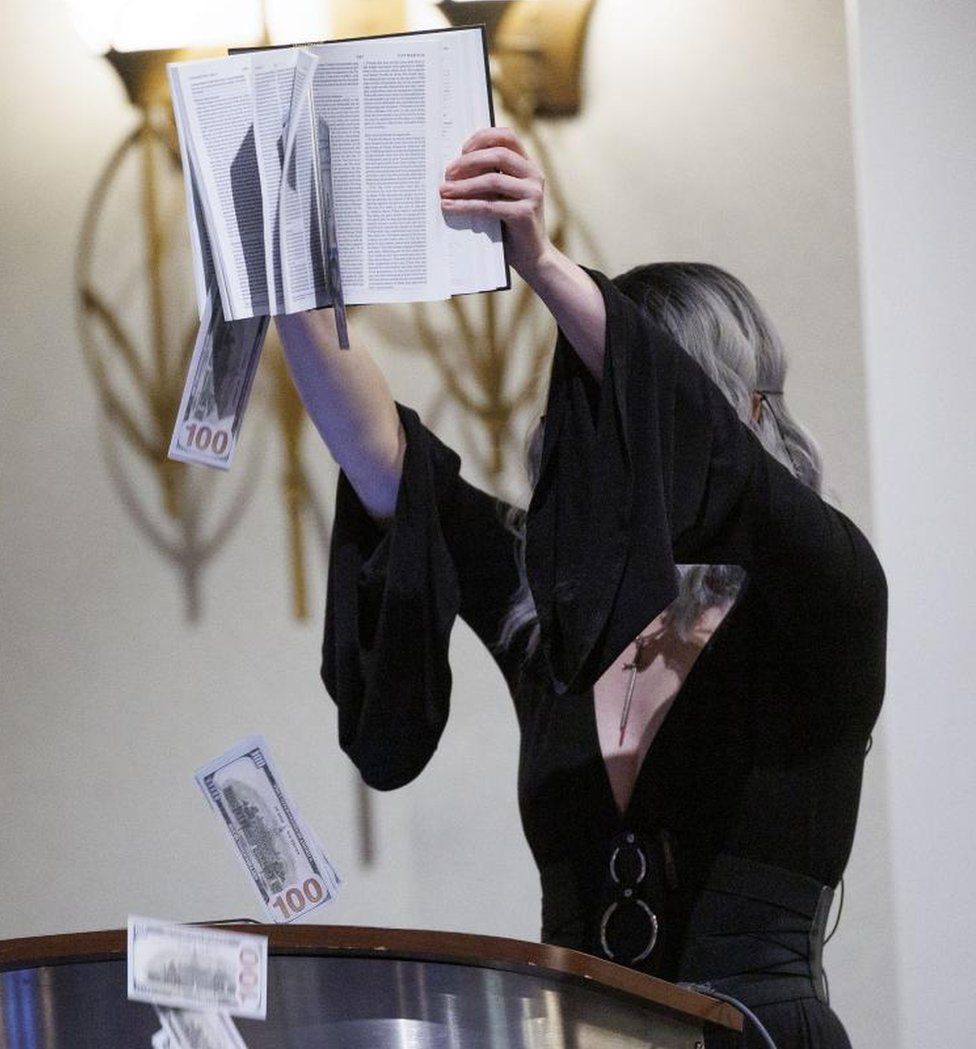 A woman in a black dress shakes fake money from a Bible during the opening statements at SatanCon 2023