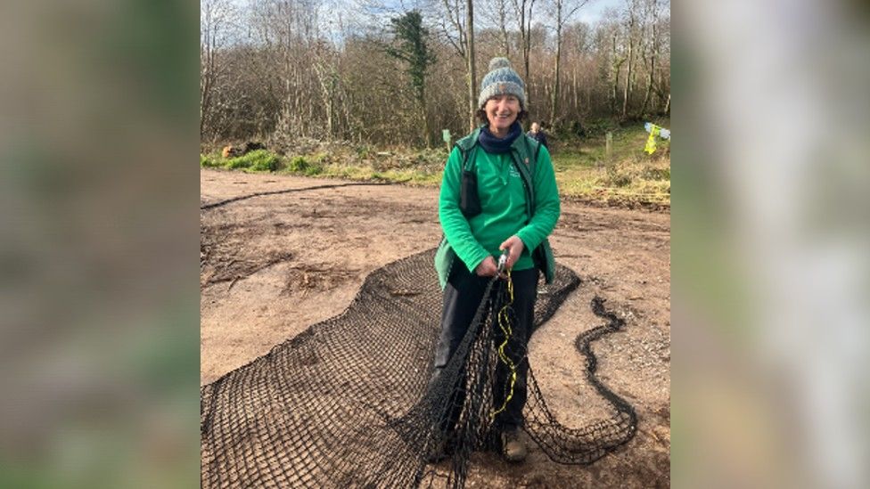 Kate Wollen - a woman in a green fleece - holding a large piece of black rope netting that will be made into a bridge for dormice