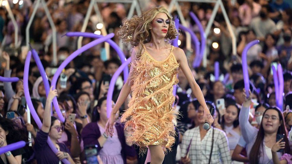 Taylor Sheesh performing to a crowd in a fringed gold slip dress and curly hair, there are purple balloons streaming in the crowd