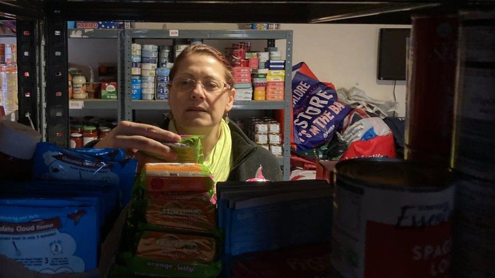 Woman with short brown hair and glasses picks up a packet of jelly from a shelf of food