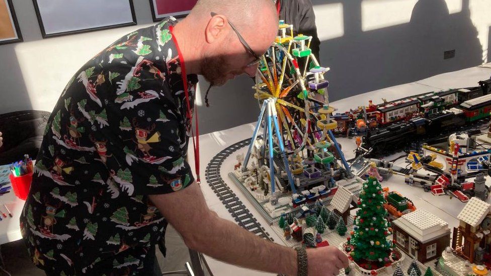 Man wearing Christmas shirt and lanyard is making adjustments to a display of lego, including a railway track and a big wheel