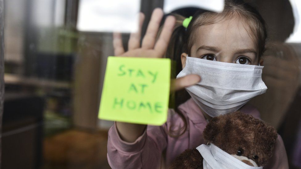 Young girl with face covering holding a teddy bear with face covering holds her hand against a window, with a note stuck to the window reading "Stay at home"