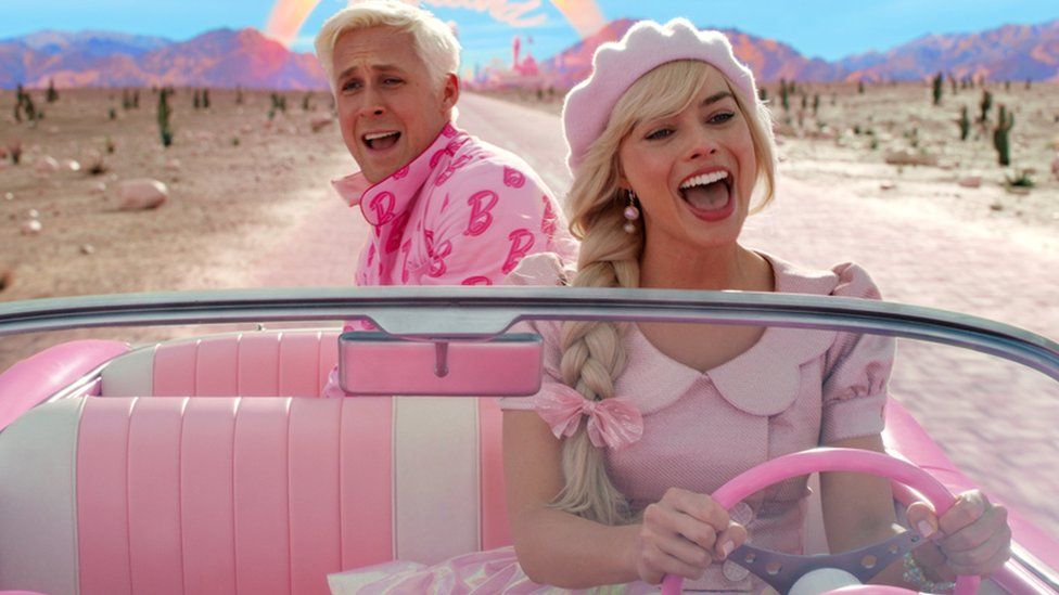 Actors Ryan Gosling and Margot Robbie in a still image taken from a scene in the Barbie movie. They're both in a pink open-top car driving along a road in what looks like a desert. A rainbow can be seen in the background over a mountain range with the words Barbieland underneath it. Margot as Barbie is wearing a pink dress and pink hat with a pink bow in her plaited blonde hair and is sat in the front seat with her hands on the pink steering wheel. Ryan as Ken is sat in the back of the car with his bleach blonde hair and a pink jacket on which is covered in the letter B. Both have their mouths open as if they are singing