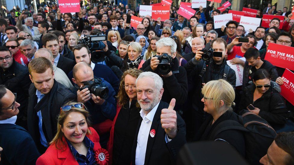 Jeremy Corbyn in the middle of a crowd, looks to the camera and raises his thumb