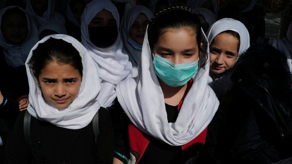 a group of primary schoolgirls, one masked, all wearing white headscarves, face the camera
