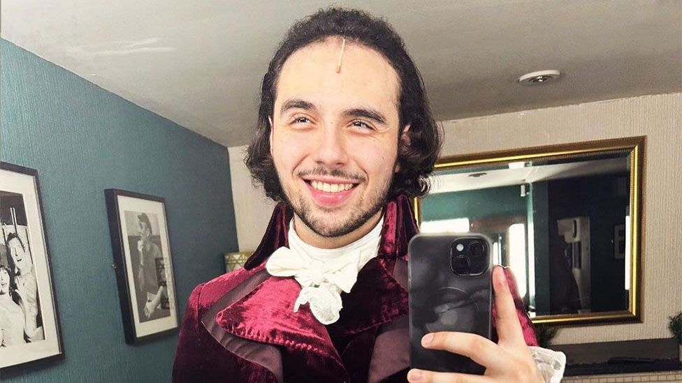 Jonathan Hermosa-Lopez taking a photo of himself in the mirror, while wearing a maroon coloured outfit set in the 1700s