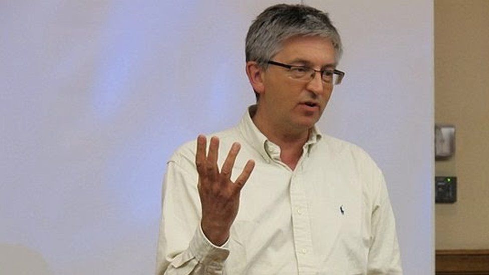 Stephen Sizer, wearing glasses and with greying hair, gesticulates as he lectures in front of a screen.
