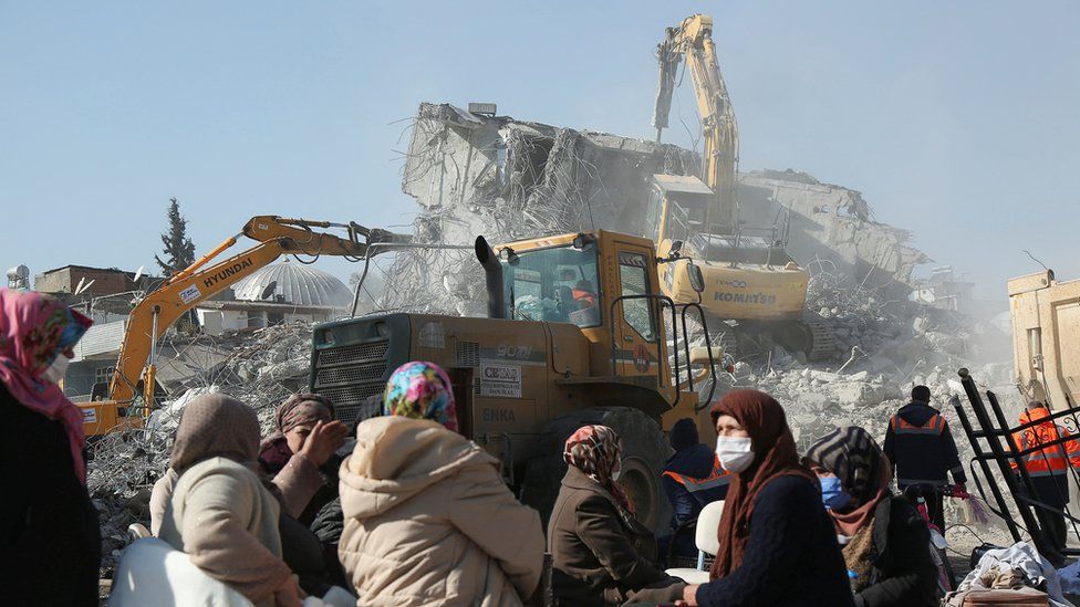 A group of people stood in front of a pile of rubble, with digger vehicles at work around the ruins of the building