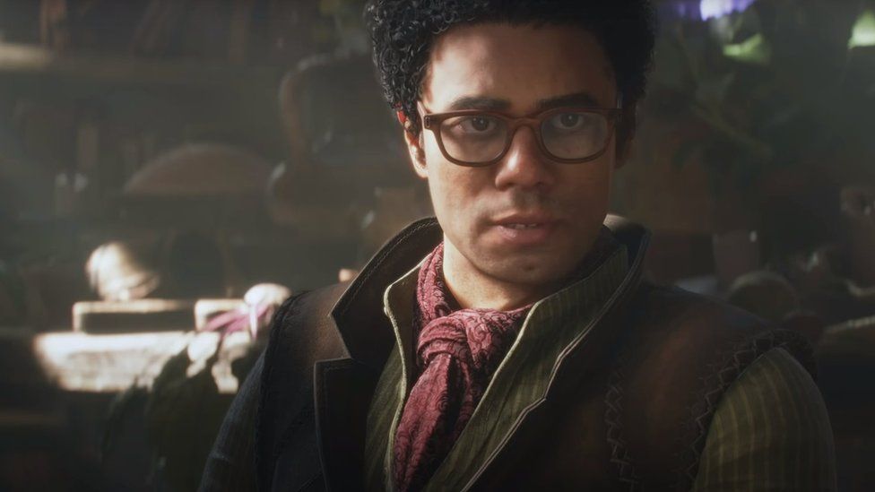 Richard Ayoade - rendered as a computer game character, speaks to the player in a screen grab of footage from Fable. He's wearing old-time clothing befitting of the game's fantasy setting - including a cravat and collarless shirt. Behind him is a dimly lit cottage containing a manner of items we can just make out the outlines of.