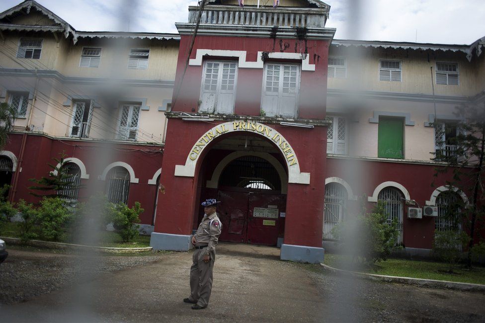 The exterior of Insein Prison, viewed through a wire fence, with a guard outside the entrance. 27 June 2016.