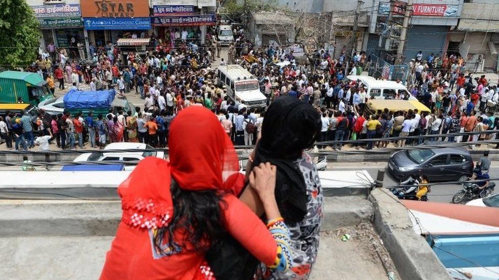Women sitting on a rooftop watch as bystanders gather while an ambulance carrying bodies of victims drives out near the site where 11 family members were found dead inside their home in the neighbourhood of Burari in New Delhi on July 1, 2018.